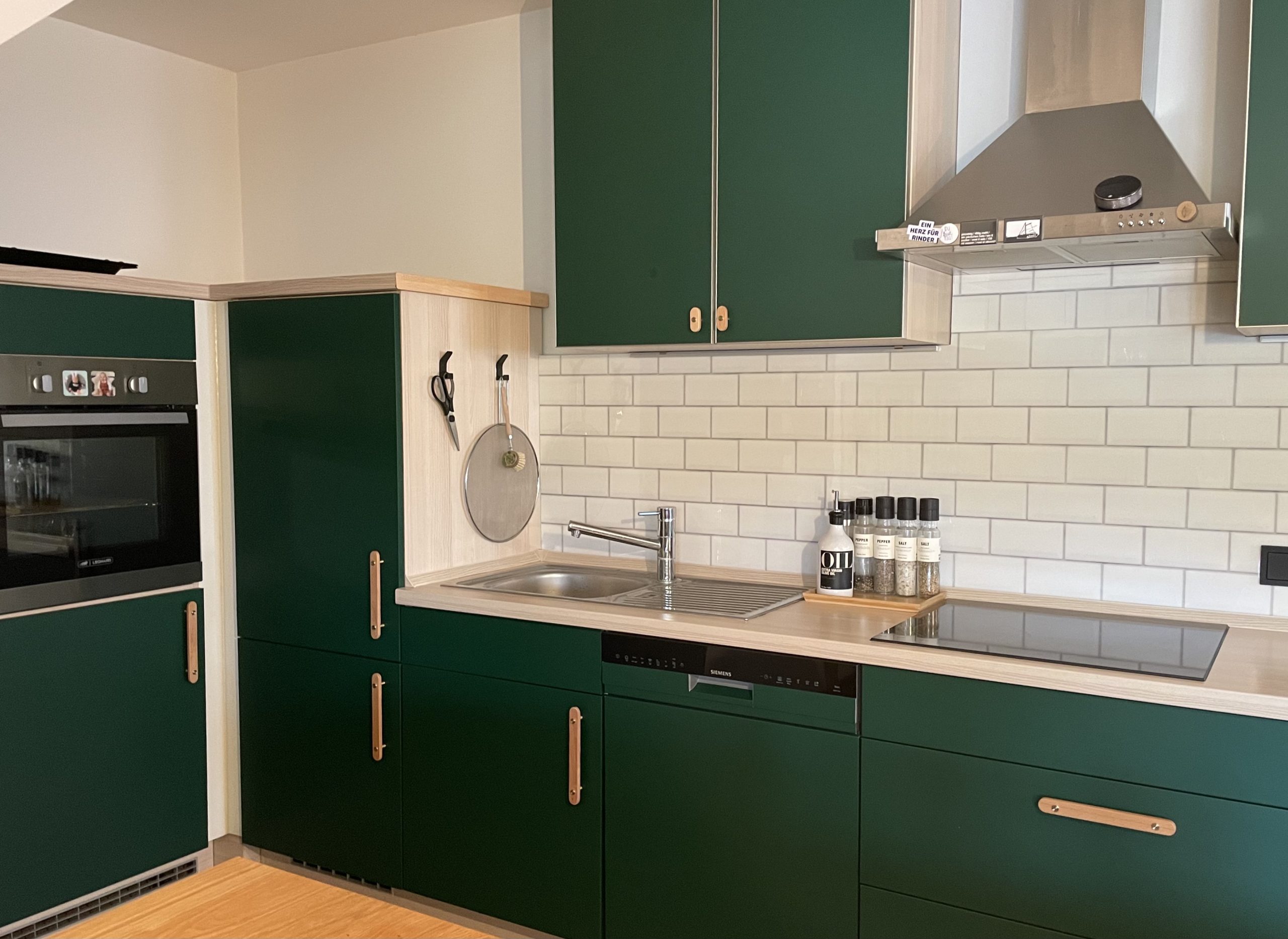Küche mit dunkelgrünen Schränken, Holzarbeitsplatte, Spüle, Herd, Dunstabzugshaube und Küchenrückwand mit weißen Kacheln