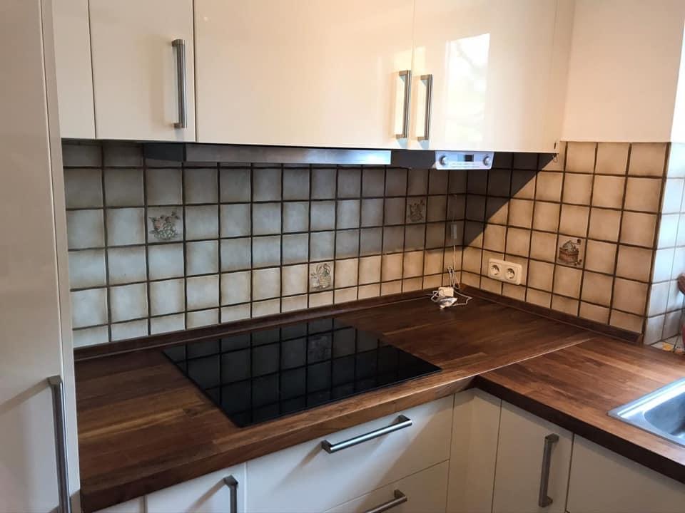 Küche mit weißen Hochglanzschränken, dunkler Holzarbeitsplatte und schwarzem Induktionskochfeld. Die Küchenrückwand ist mit kleinen, grau-beigen quadratischen Fliesen und vereinzelten Dekorfliesen gestaltet. Rechts befindet sich eine Edelstahlspüle.