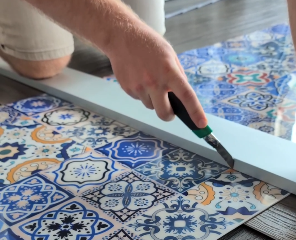 Hand schneidet mit einem Teppichmesser entlang einer Leiste eine Küchenrückwand mit einer marokkanisch inspirierten Fliesenoptik zu.